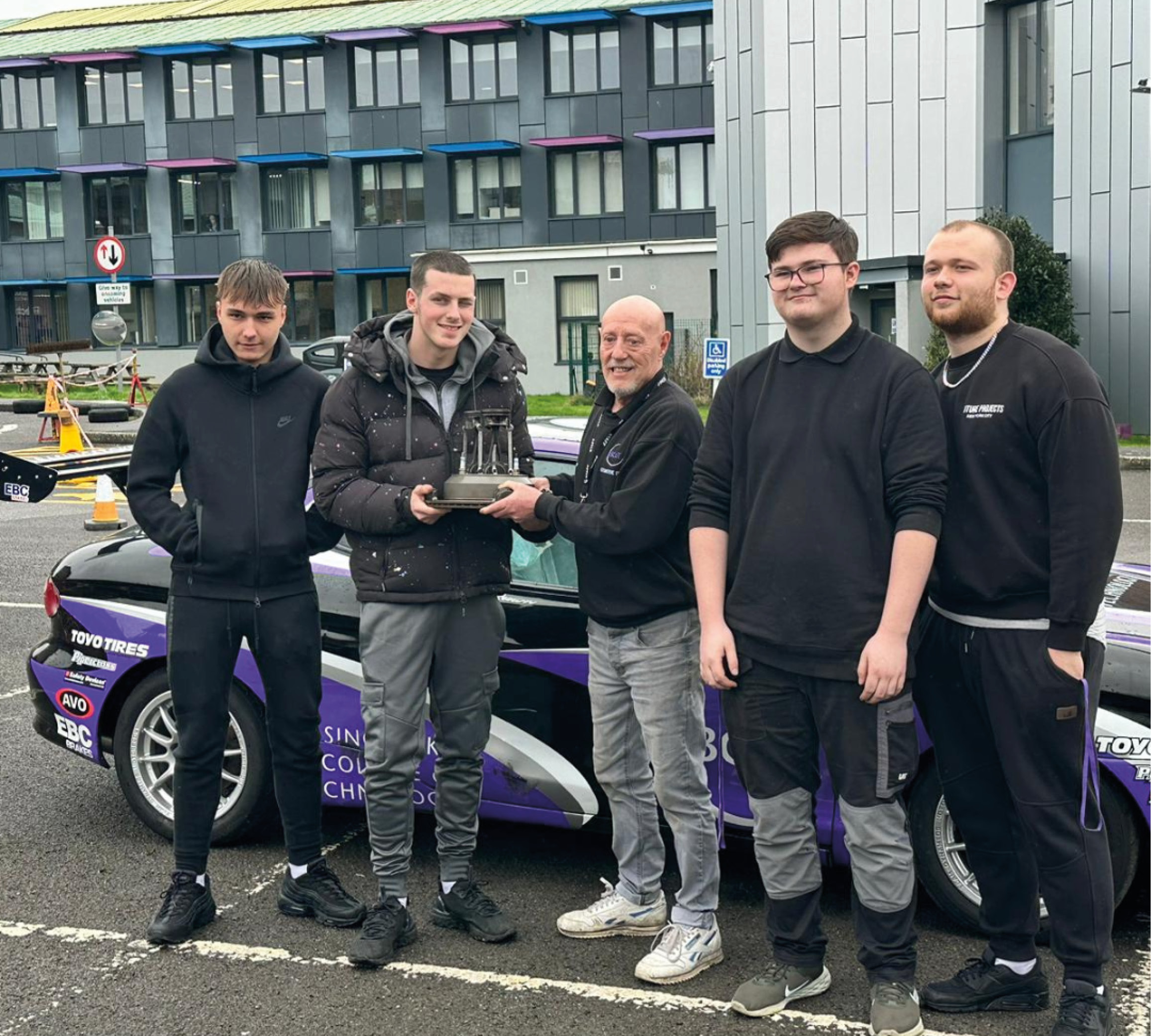 Students standing in front of racing car being handed trophy by teacher